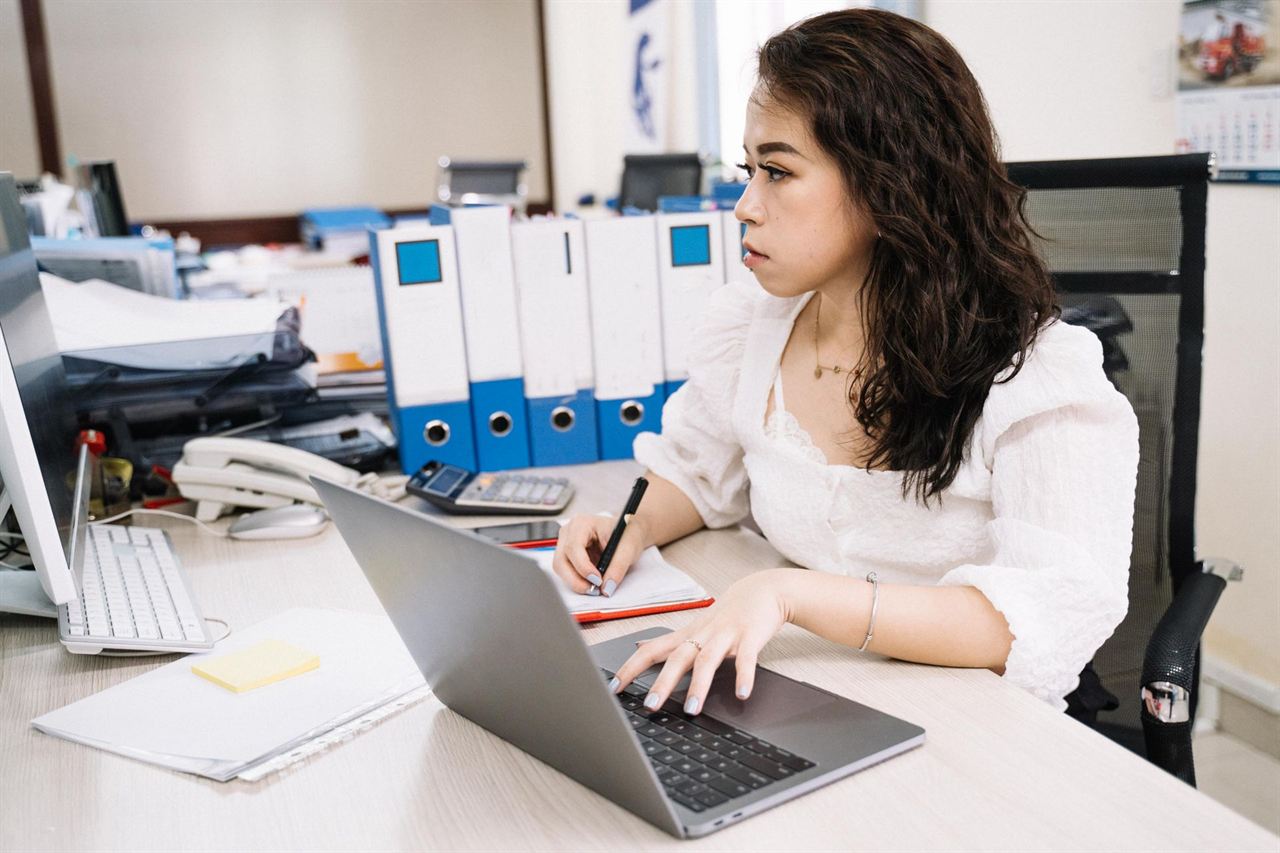 書類のファイルが並ぶ事務所で、ノートパソコンに向かって仕事をする女性